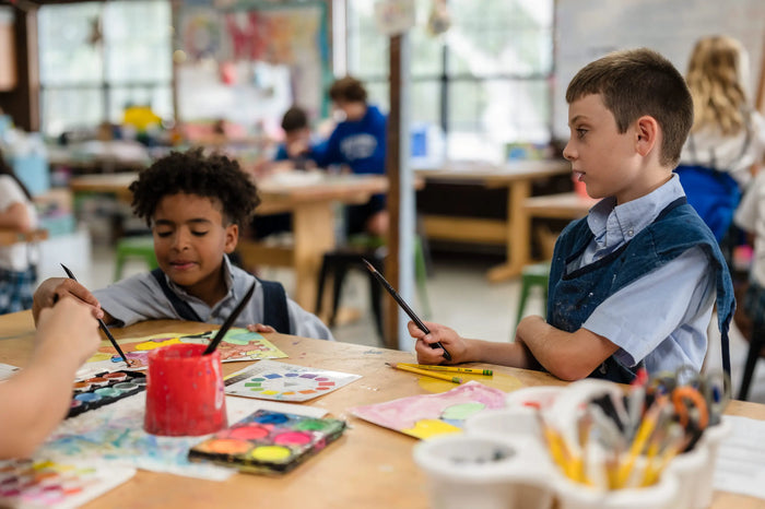 Elementary school students in Kehoe-France uniforms painting together in an art classroom, sitting at a table with watercolor supplies and brushes.