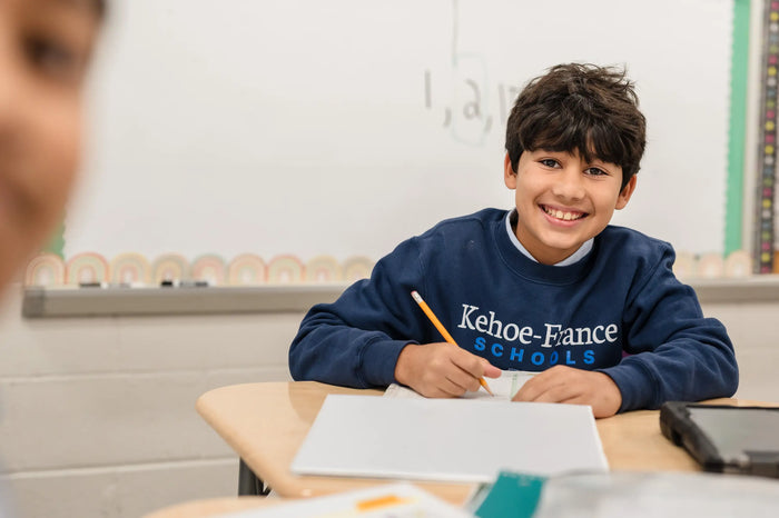 Middle school student in a Kehoe-France uniform sweatshirt smiling while writing at his desk in a classroom.
